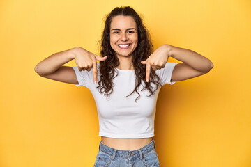 Young Caucasian woman, yellow studio background, points down with fingers, positive feeling.