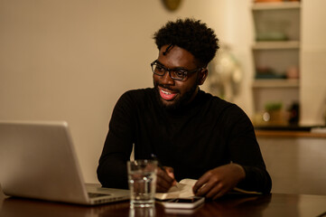 A cheerful multicultural man is sitting at home at night and following a webinar on a laptop while smiling at it.