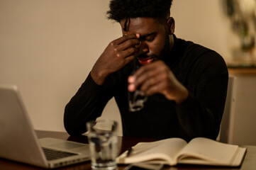 An interracial entrepreneur is having a headache while sitting at a table at home surrounded by a laptop and textbook.