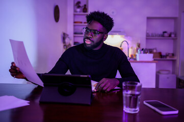 A focused interracial man is sitting in a purple lighted room at home and doing paperwork and home...