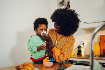 A caring african american mother is blowing her son's nose in a kitchen at home.