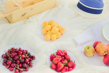 Fototapeta na wymiar Summer fruits, apricots, strawberries, cherries, apples, empty wine glass, wooden box, hat on beige blanket.