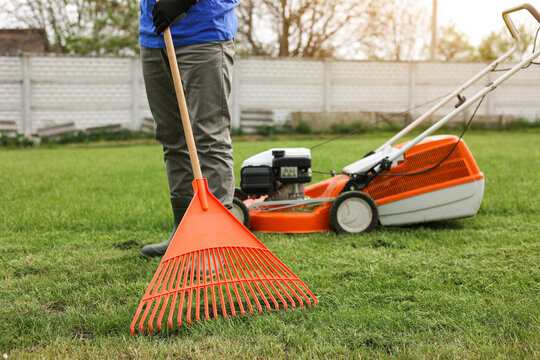 Male gardener collects cut grass with orange plastic rake after a mower, works in the backyard of the house. Man takes care of lawn. Concept of housework, gardening and country life, garden tools.