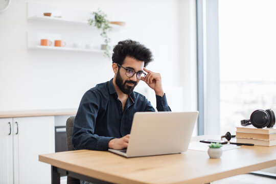 Confident indian man in dark collared shirt using laptop while sitting at writing desk on kitchen background. Efficient freelancer searching information while doing full-time job in home interior.