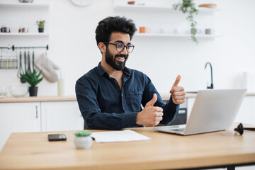 Fototapeta premium Cheerful bespectacled adult waving hello at webcam of portable computer while greeting colleague in home office. Proficient indian consultant receiving online conference call using technologies.