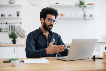 Serious young man in business casual clothes holding online conference with coworkers from home office. Focused indian entrepreneur discussing company issues using portable computer in apartment.