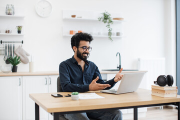 Fototapeta premium Mindful arabian male in business casual clothes using microphone of headset while holding online briefing via laptop. Efficient remote employee informing colleagues about working arrangement at home.