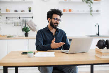 Mindful arabian male in business casual clothes using microphone of headset while holding online briefing via laptop. Efficient remote employee informing colleagues about working arrangement at home.