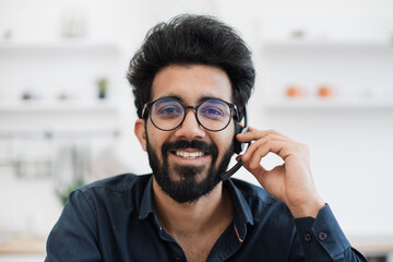 Close up view of indian man holding microphone of hands-free headset while smiling at camera on kitchen background. Happy professional working virtually as staff member for fully remote company.