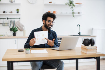 Fototapeta premium Attractive indian male holding paper with charts while speaking during online meeting via computer at home. Qualified data analyst demonstrating information about business growth in kitchen interior.