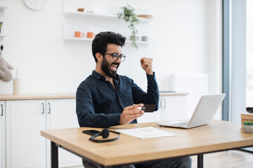 Fototapeta premium Emotional bespectacled adult with debit card lifting fist in excitement while looking happily at computer screen in kitchen. Delighted indian male making successful purchase using internet banking.