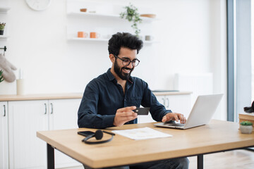 Fototapeta premium Joyful arabian man in casual clothes using black credit card while running portable computer in workplace. Smiling bearded person in glasses enjoying virtual shopping using online wallet from home.