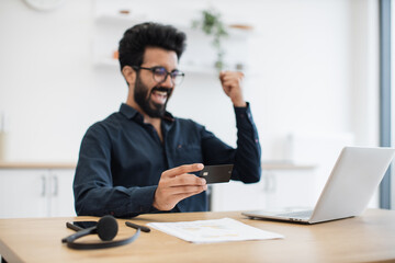 Emotional bespectacled adult with debit card lifting fist in excitement while looking happily at computer screen in kitchen. Delighted indian male making successful purchase using internet banking.