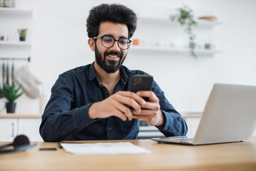 Positive indian male in spectacles holding smartphone while staying in modern workplace at home. Efficient freelance worker texting message to colleague while performing project for employer indoors.