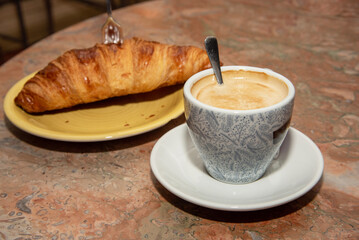 Close-up of a delicious, shiny croissant with a cup of coffee and milk on a weathered wooden table.