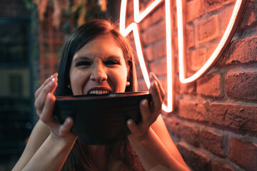 Amused young woman holding her poke bowl in her hands and biting it aggressively. Focus on the girl.