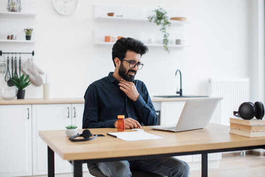 Sick young man in business casual clothes touching sore throat while leveraging modern laptop in dining room. Deseased arabian employee working remotely from home while fighting illness indoors.