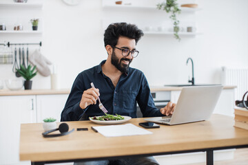 Obraz na płótnie Canvas Smiling mature man in business casual clothes eating healthy salad while looking at laptop screen in kitchen interior. Indian remote employee having lunch break while doing home-based job at midday.