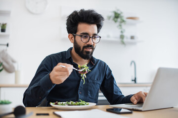 Efficient indian worker in glasses eating vegetables while typing on portable computer at wooden desk in kitchen. Remote freelancer performing business task without wasting time for lunch break.
