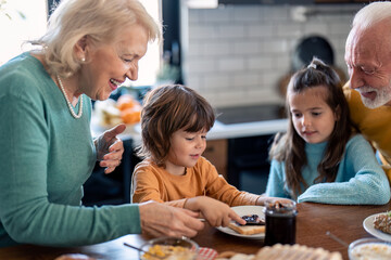 Small boy enjoying breakfast with sister and grandparents in the kitchen at home. Happy family preparing morning meal at dining table in the kitchen.