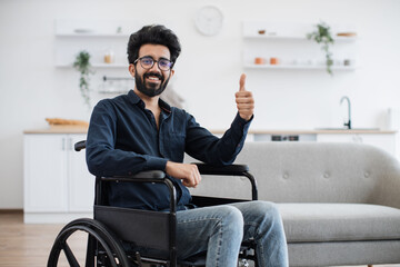 Full length portrait of arabian wheelchair user smiling at camera while resting in open-plan kitchen of modern apartment. Cheerful young male in casual clothes and glasses spending pastime indoors.