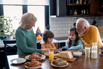 Happy kids enjoying breakfast with senior woman and senior man - grandparents in the kitchen at home. Happy family preparing morning meal at dining table in the kitchen.