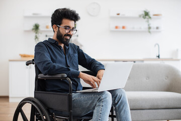 Joyful indian man in wheelchair holding laptop on knees while staying in middle of bright open-plan kitchen. Happy young adult surfing internet webpages while recovering after accident at home.