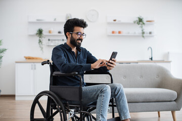 Smiling arabian adult in wheelchair looking at cell phone screen while having rest in comfortable...