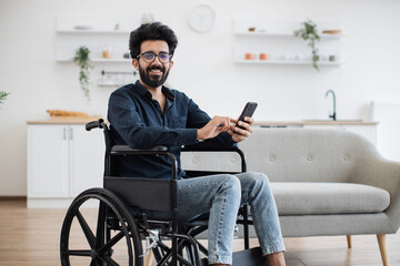 Smiling arabian adult in wheelchair looking at cell phone screen while having rest in comfortable dining room. Cheerful positive man checking social media using internet connection in apartment.
