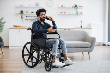 Full length view of adult person in wheelchair talking on cell phone while using comfort of modern studio flat. Mature arabian man organising birthday party while getting recovery from illness.