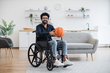 Full length portrait of indian adult with mobility issues holding ball in hands before doing daily exercises at home. Wheelchair basketball player in casual clothes staying positive in room interior.