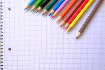 school background, colored pencils lie on a sheet of paper, an open notebook on a spiral, a college block in a cage, close-up, the concept of student writing supplies, back to school