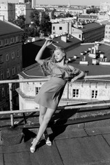 Obraz premium Elegant young lady in ruffle attire, heels shoes stand on rooftop leaning on fence with closed eyes and lifting arm. 