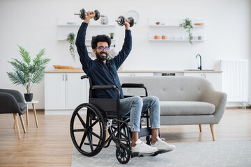 Joyful arabian person with physical disability holding dumbbells in raised arms during strength...