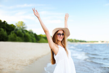Happy smiling woman in free happiness bliss on ocean beach standing with a hat, sunglasses, and rasing hands. Portrait of a multicultural female model in white summer dress enjoying nature during
