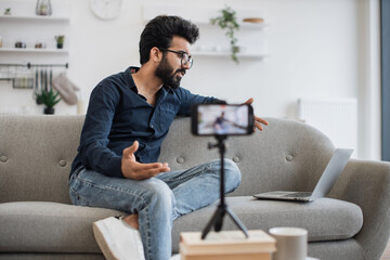 Indian man with stylish haircut reading information from modern laptop during live broadcast on social network. Demanding tutor using modern smartphone with tripod for online training.