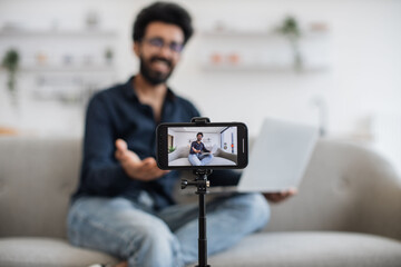 Blurred background of cheerful arabian man sitting on couch with laptop and talking something on...