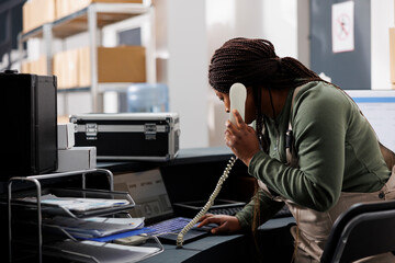 Stockroom supervisor talking at landline phone with remote client, discussing orders details in...