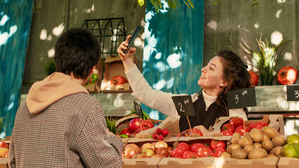 Young farmer recording video on smartphone with regular client, promoting small farming business on social media. Female vendor filming with mobile phone, having fun with customer.