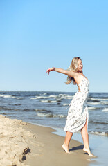 Fototapeta na wymiar Happy blonde beautiful woman on the ocean beach standing in a white summer dress, open arms