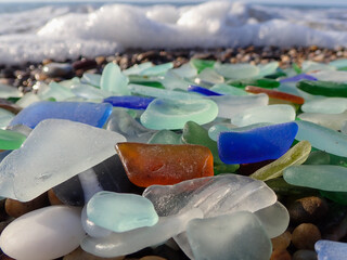 Sea glass, natural polish textured sea glass and stones on the seashore. Azure clear sea water with waves. Green, blue shiny glass with multi-colored sea pebbles close-up. Beach summer background.