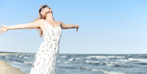 Happy blonde beautiful woman on the ocean beach standing in a white summer dress and sun glasses, open arms