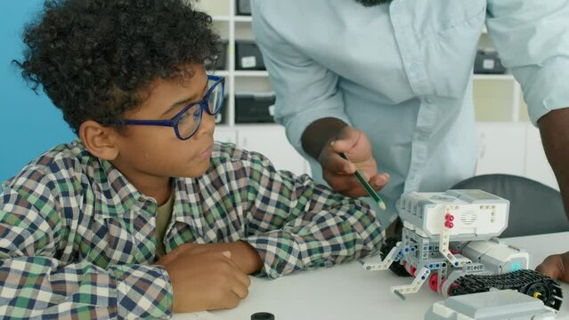 Medium close-up tilting shot of young African American male technology teacher talking to preteen black boy in glasses in after school club and explaining design and function of model robot