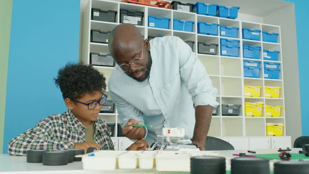 Medium shot of 9-year-old curly African American boy in glasses and gingham shirt sitting at table in kids after school club and building robot model, and black male teacher giving him advice