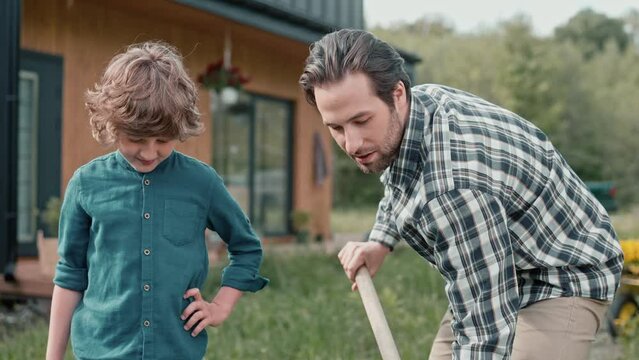 Young boy watching his dad working with gardening tools. Busy father communicating with his son while planting trees or plants. Working together outside. Teamworking. Concept of relationship.