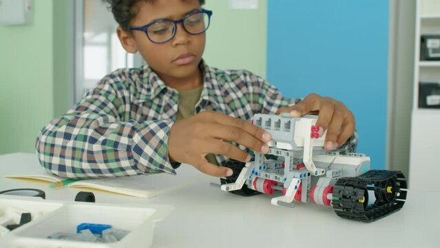 Medium close-up shot of nerdy 9-year-old African American male child in glasses and plaid shirt sitting at table in after school hobby club and making toy robot from plastic blocks