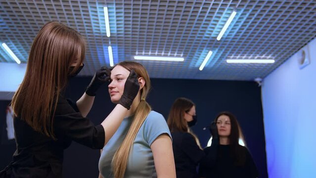 Brow master marking the lines of girl's eyebrows with the thread. Beautician draws lines with white pen to a woman at backdrop.