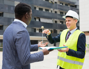 European man architect giving keys to apartment in new building to African-american man while standing outdoors.