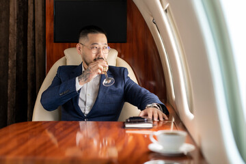 successful asian businessman in suit and glasses flies in private jet drinks champagne and looks out the window