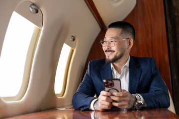 successful asian businessman in suit and glasses flies in private luxury jet and uses smartphone